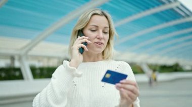 Parkta elinde kredi kartıyla akıllı telefondan konuşan genç sarışın kadın.