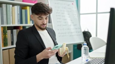 Genç Arap erkek öğrenci üniversite sınıfında akıllı telefon kullanarak muz yiyor.