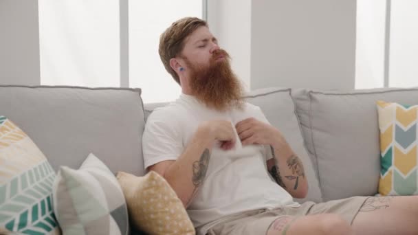 年轻的红头发男子坐在沙发上 在家里汗流浃背 — 图库视频影像