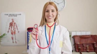 Klinikte steteskop tutan genç sarışın doktor gülümsüyor.
