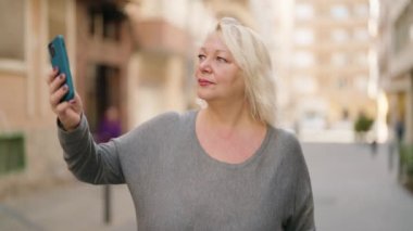 Orta yaşlı sarışın kadın gülümsüyor. Kendine güveni tam. Sokakta akıllı telefonun yanında selfie çekiyor.