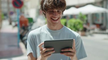 Young blond man smiling confident watching video on touchpad at street
