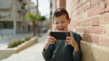 Sarışın çocuk sokakta akıllı telefondan video izliyor.