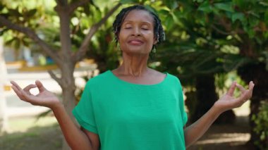 Orta yaşlı Afro-Amerikan kadın parkta yoga egzersizi yaparken gülümsüyor.