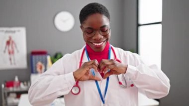 Afrikalı Amerikalı kadın doktor gülümsüyor klinikte elleriyle kalp hareketi yapıyor.