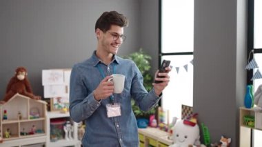 Young hispanic man preschool teacher using smartphone drinking coffee at kindergarten