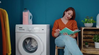 Çamaşır odasında kitap okuyan genç kızıl saçlı kadın çamaşır makinesini bekliyor.