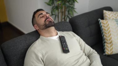 Televizyon kumandalı genç İspanyol adam kanepede oturup evde uyuyor.