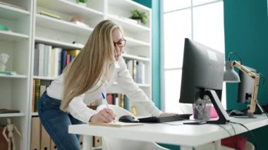 Young blonde woman student using computer writing on notebook at library university