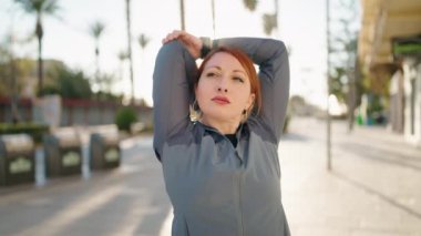 Genç kızıl saçlı kadın spor giyer sokakta esneme hareketleri yapar.