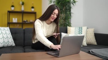 Young beautiful hispanic woman writing on notebook using laptop at home