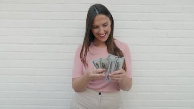 Young beautiful hispanic woman smiling confident counting dollars over isolated white brick background