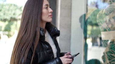 Genç, güzel, İspanyol bir kadın akıllı telefon kullanıyor. Sokakta giyim mağazası bakıyor.
