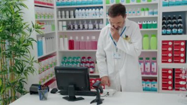 Middle age man pharmacist talking on telephone using computer at pharmacy