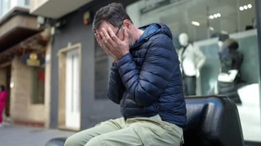 Orta yaşlı bir adam sokakta endişeli bir ifadeyle bankta oturuyor.