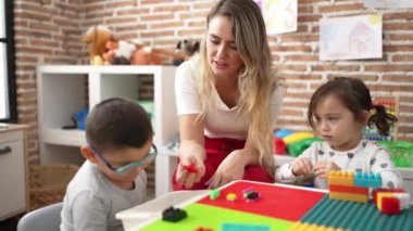 Teacher with boy and girl playing with construction blocks sitting on table at kindergarten