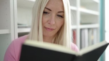 Young blonde woman teacher reading book doing silence gesture at library university