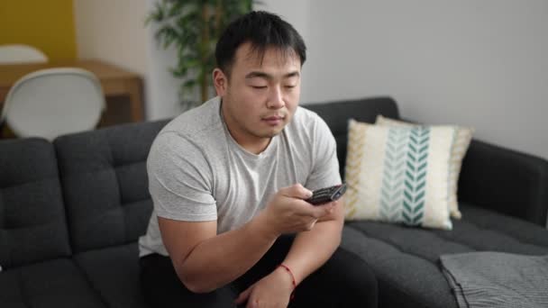 Молодой Китаец Смотрит Телевизор Сидя Диване Скучным Выражением Лица Дома — стоковое видео