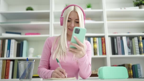 在图书馆大学 年轻的金发女学生用智能手机和耳机在笔记本上写字 — 图库视频影像