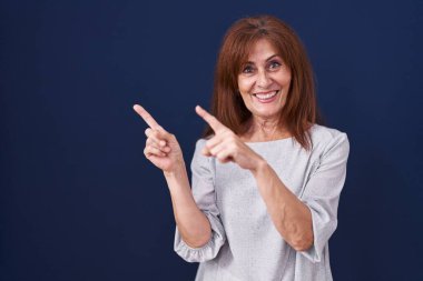 Orta yaşlı bir kadın mavi arka planda duruyor gülümsüyor ve kameraya bakıyor iki eliyle ve parmaklarıyla kenarı işaret ediyor.. 