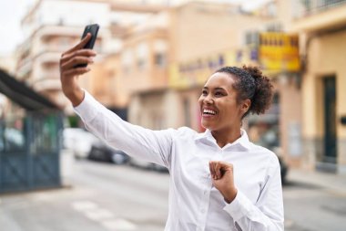 Afro-Amerikalı kadın gülümsüyor. Sokakta akıllı telefonun yanında özçekim yapıyor.