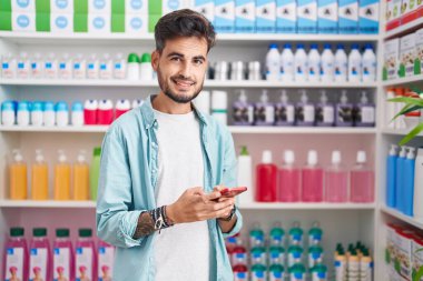 Genç İspanyol adam eczanede akıllı telefon kullanarak gülümsüyor.