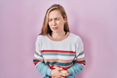 Young blonde woman standing over pink background with hand on stomach because indigestion, painful illness feeling unwell. ache concept. 