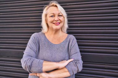 Orta yaşlı sarışın kadın gülümsüyor. Kollarını kavuşturup caddede duruyor.