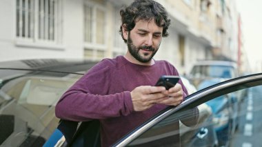Young hispanic man using smartphone leaning on car at street