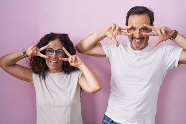 Orta yaşlı İspanyol çift birlikte pembe arka planda barış sembolü yapıyorlar. Yüzlerinde parmakları, gülümseyerek zafer gösteriyorlar. 