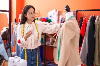 Young chinese woman tailor holding notebook standing by manikin at atelier