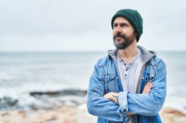 Kel genç adam sahilde kollarını kavuşturmuş el kol hareketi yapıyor.