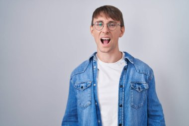 Caucasian blond man standing wearing glasses angry and mad screaming frustrated and furious, shouting with anger. rage and aggressive concept. 