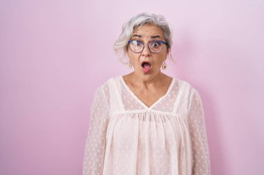 Pembe arka planda duran gri saçlı orta yaşlı bir kadın. Şaşırmış ve korkmuş bir ifade, korku ve heyecanlı bir yüz ifadesi var.. 