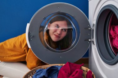 Genç esmer kadın çamaşır makinesinin camından bakıyor gülümsüyor yan tarafa bakıyor ve düşünüyor.. 