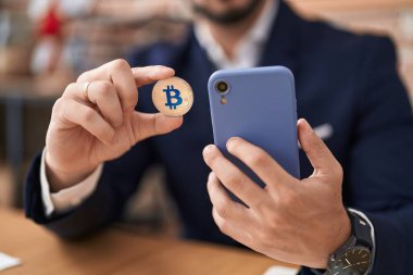 Genç İspanyol iş adamı ofisinde Bitcoin tutarak akıllı telefon kullanıyor.