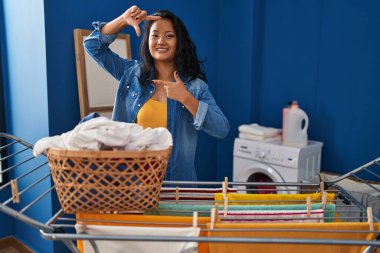 Genç Asyalı kadın çamaşır ipine kıyafet asıyor gülümsüyor ve elleri ve parmakları gülen yüzlü bir çerçeve yapıyor. yaratıcılık ve fotoğrafçılık kavramı. 