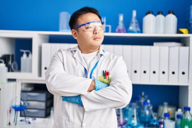 Bilim adamı üniforması giyen genç Çinli adam laboratuarda kollarını kavuşturup el kol hareketi yapıyor.