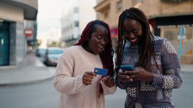 İki Afro-Amerikalı arkadaş sokakta akıllı telefon ve kredi kartı kullanarak gülümsüyor.