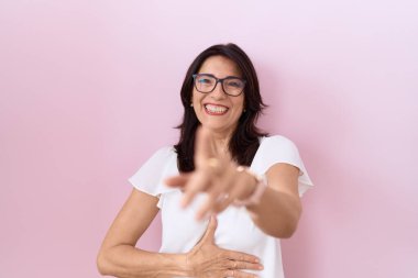 Orta yaşlı, beyaz tişörtlü ve gözlüklü bir İspanyol kadın sana gülüyor, eli vücudunun üzerinde kamerayı işaret ediyor, yüz ifadesi utanç verici. 