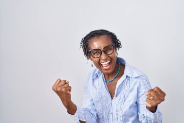 Beyaz arka plan üzerinde duran Afrikalı kadın gözlük takıyor. Çok mutlu ve heyecanlı. Kolları havada, gülümseyen ve başarı için çığlık atan kazanan bir jest yapıyor. kutlama konsepti. 