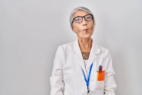 Middle age woman with grey hair wearing scientist robe making fish face with lips, crazy and comical gesture. funny expression.