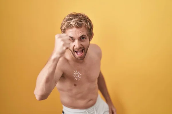 Caucasian man standing shirtless wearing sun screen angry and mad raising fist frustrated and furious while shouting with anger. rage and aggressive concept.