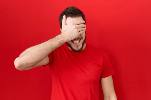 カジュアルな赤いTシャツを着た若いヒスパニック系の男性が笑顔で 驚きのために目を覆う顔で手で笑っています 視覚障害の概念 — ストック写真