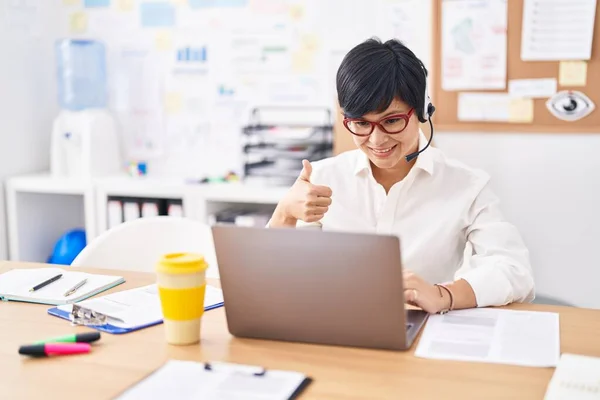 stock image Young asian woman with short hair wearing call center agent headset smiling happy and positive, thumb up doing excellent and approval sign 