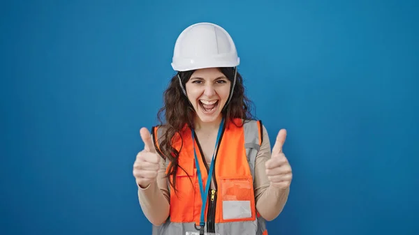 年轻美丽的惊慌失措的女建筑工人带着自信的微笑 在孤独的蓝色背景下竖起大拇指做着手势 — 图库照片
