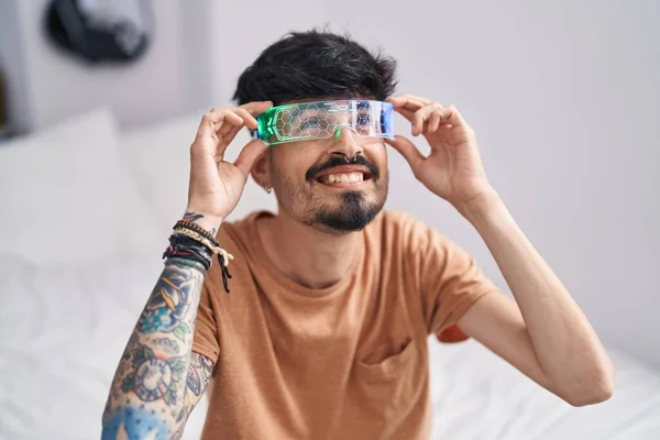 stock image Young hispanic man using augmented reality glasses sitting on bed at bedroom