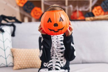 İskelet kostümlü sevimli İspanyol çocuk evde balkabağı sepeti tutuyor.