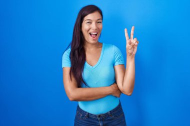Young hispanic woman standing over blue background smiling with happy face winking at the camera doing victory sign. number two. 