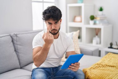 Hispanic man with beard using touchpad sitting on the sofa showing middle finger, impolite and rude fuck off expression 